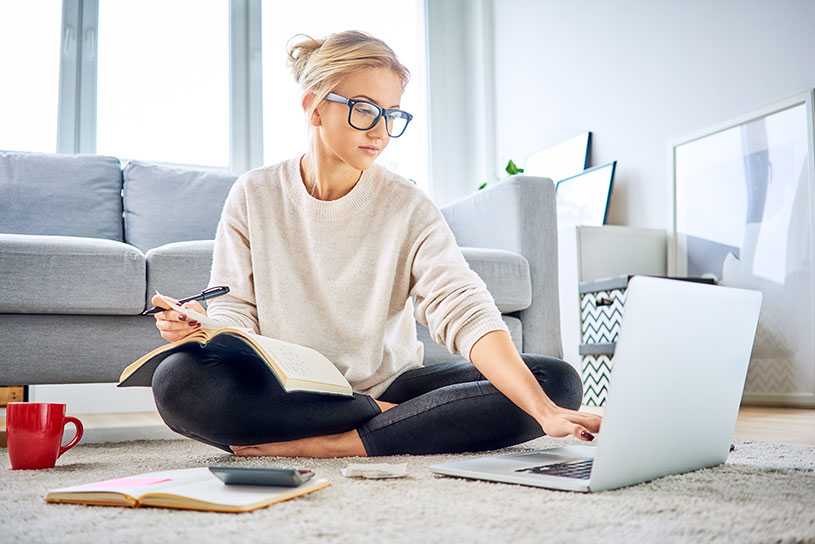 Woman sat on the floor in front of sofa, wearing glasses and using a laptop to pay bills online