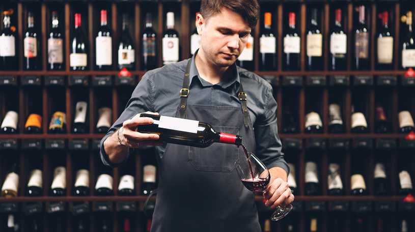 Wine bartender pouring a glass of red wine in front of shelves of wine