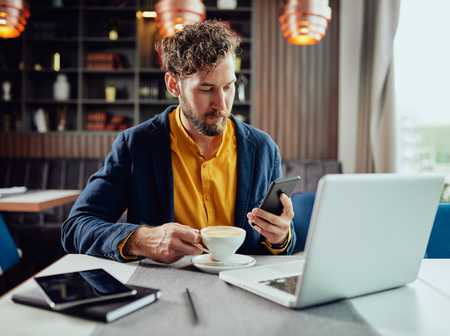 Man using mobile phone and drinking coffee in front of laptop