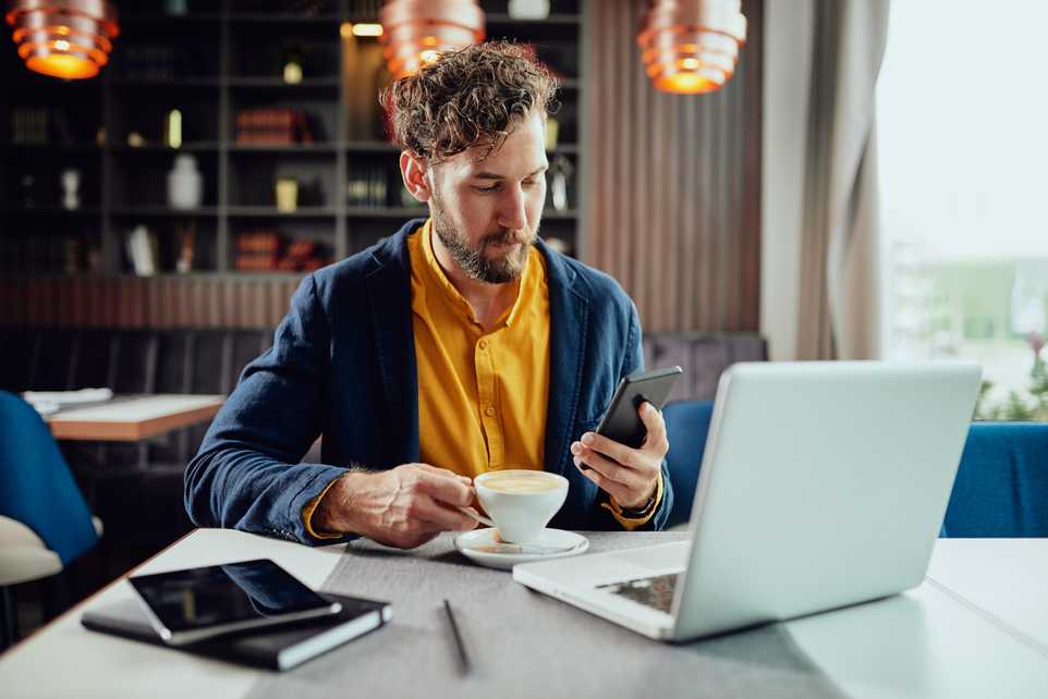 Man using mobile phone and drinking coffee in front of laptop
