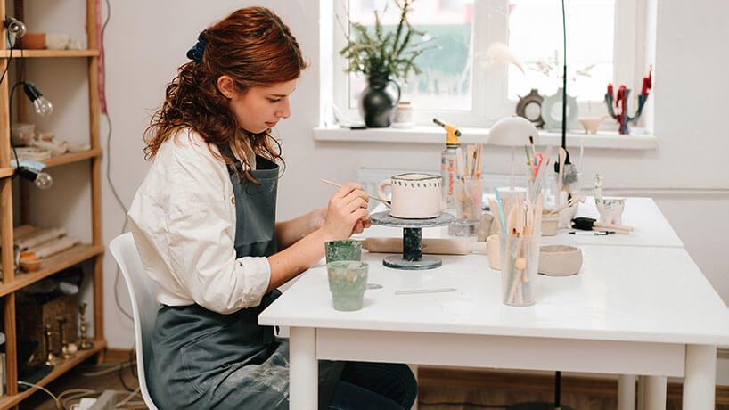 Artist is decorating and painting handmade ceramic clay cup in the creative workshop. Painter is drawing and adding unique designs to the earthenware.