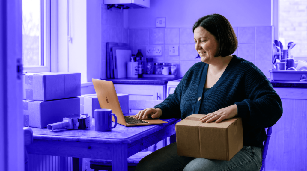A shop owner works on a laptop and holds a package for her small business