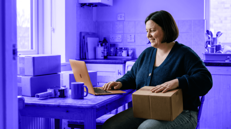 A shop owner works on a laptop and holds a package for her small business