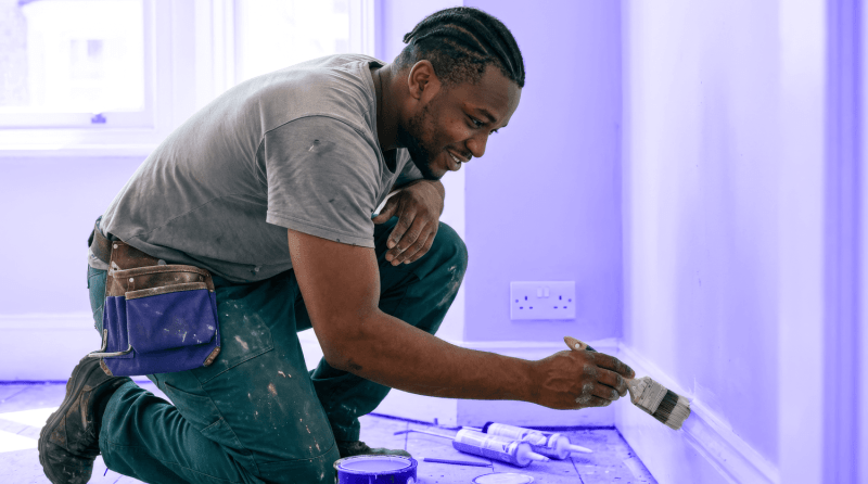 A landlord does landlord maintenance on his rental property, kneeling down and painting a skirting board