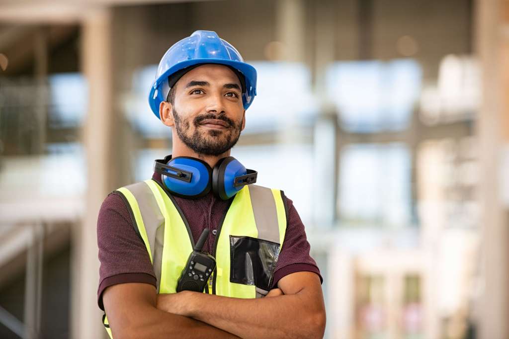 Small business owner wearing PPE including blue hat crosses arms