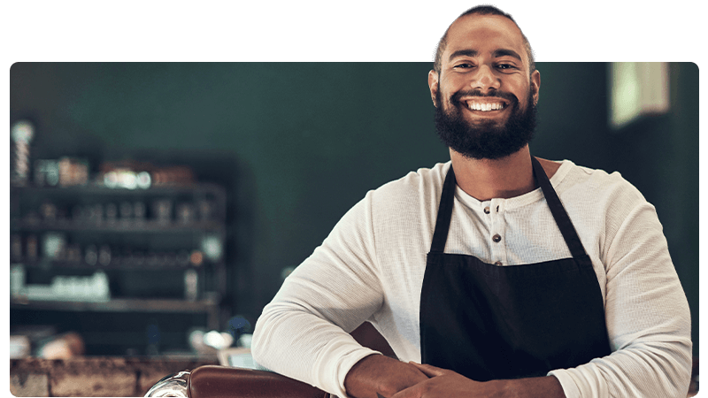 Barber in an apron smiles at the camera and leans on his chair.