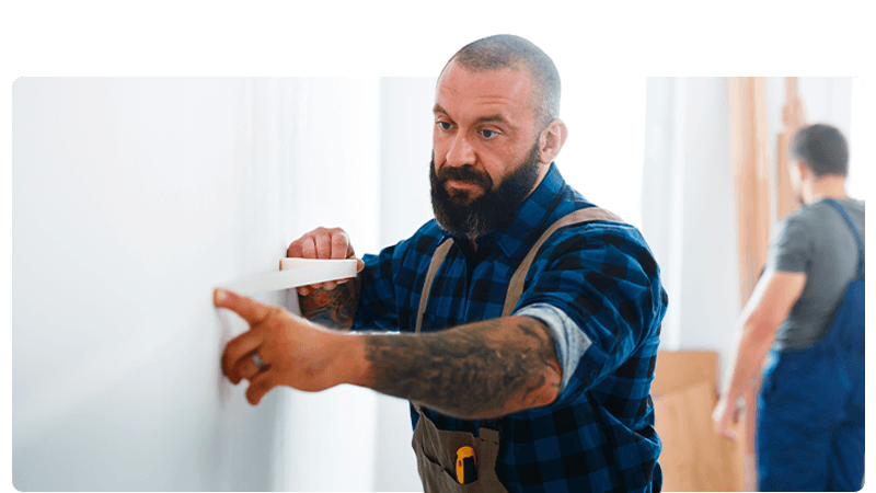 Bedroom fitter in overalls and a blue shirt masking a wall while a man works behind him.