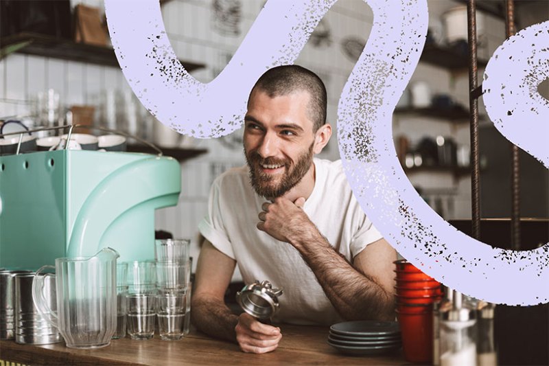 A male barista smiles while leaning on the counter of a coffee shop
