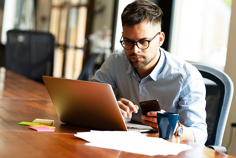 Serious man in glasses using laptop and phone with paperwork
