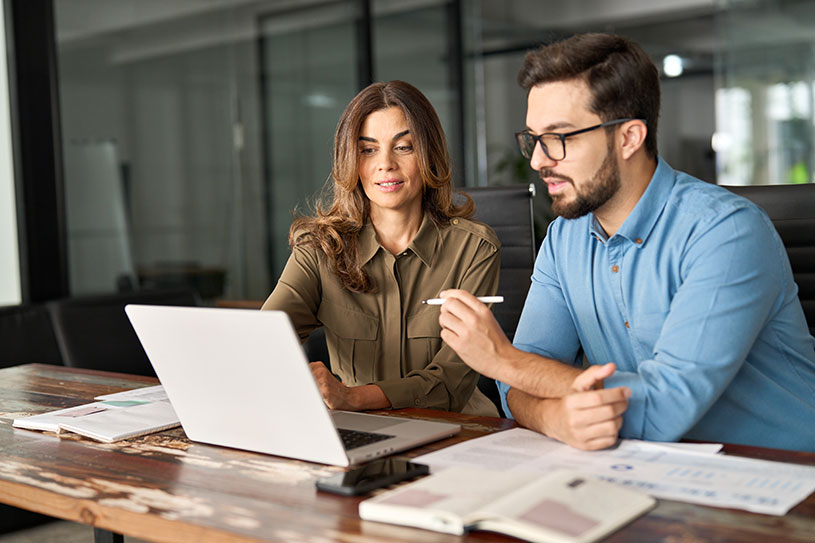 Two people working in an office, looking at a computer and taking notes