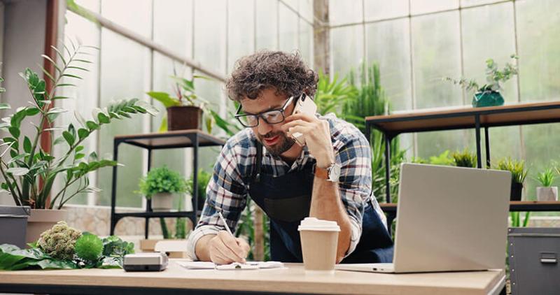 Happy Caucasian businessman talking on cellphone while standing in apron in small floral center and writing down order details.