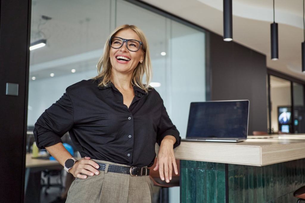 A confident, smiling businesswoman with stylish glasses radiates positivity at a modern, sleek office workspace