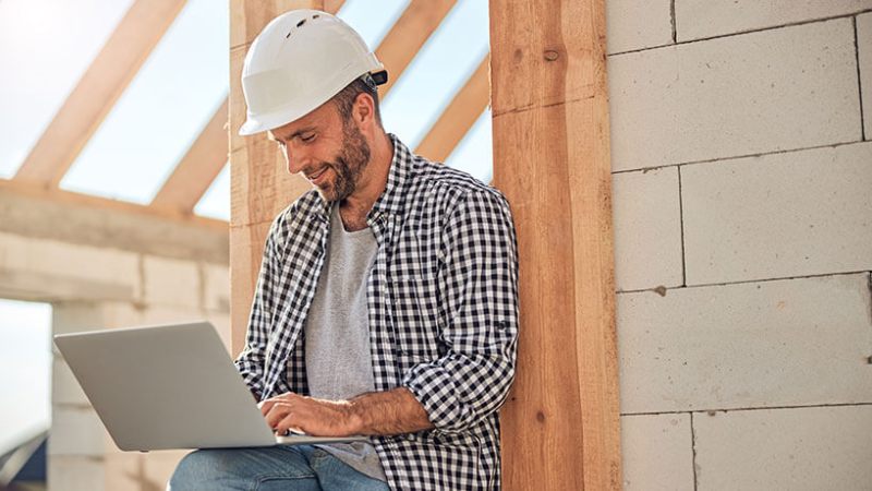 A male builder in a hard hat works on a laptop while on a building site