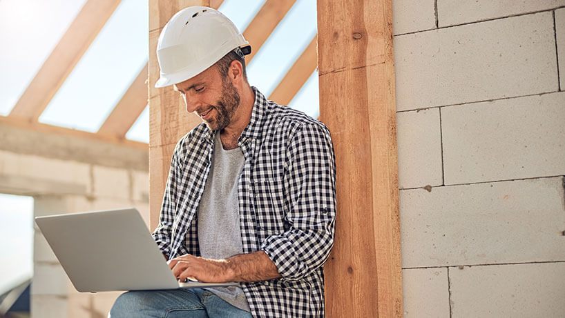 A male builder in a hard hat works on a laptop while on a building site