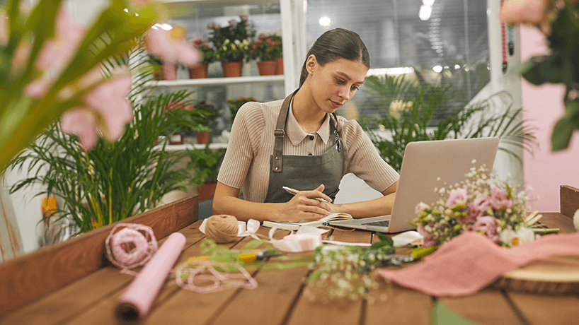 Portrait of female small business owner managing accounting books while using laptop at table in flower shop, copy space